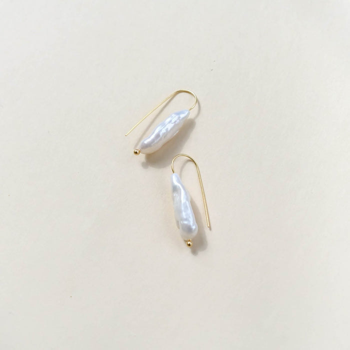 The Here to Stay Earrings by LLUME Jewelry feature irregular freshwater white pearls on gold plated hooks, shown against a light beige background.