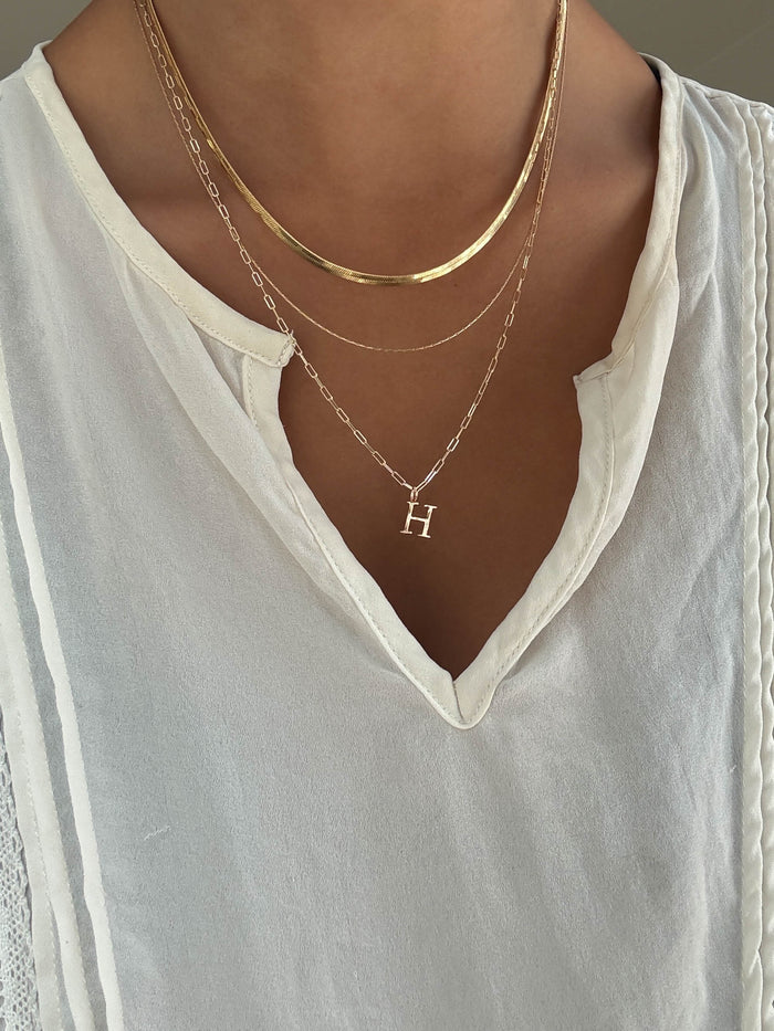 A person wears a white V-neck blouse and LLUME Jewelry's Initial Necklace Set—three layered chains, one with an 'H' pendant.