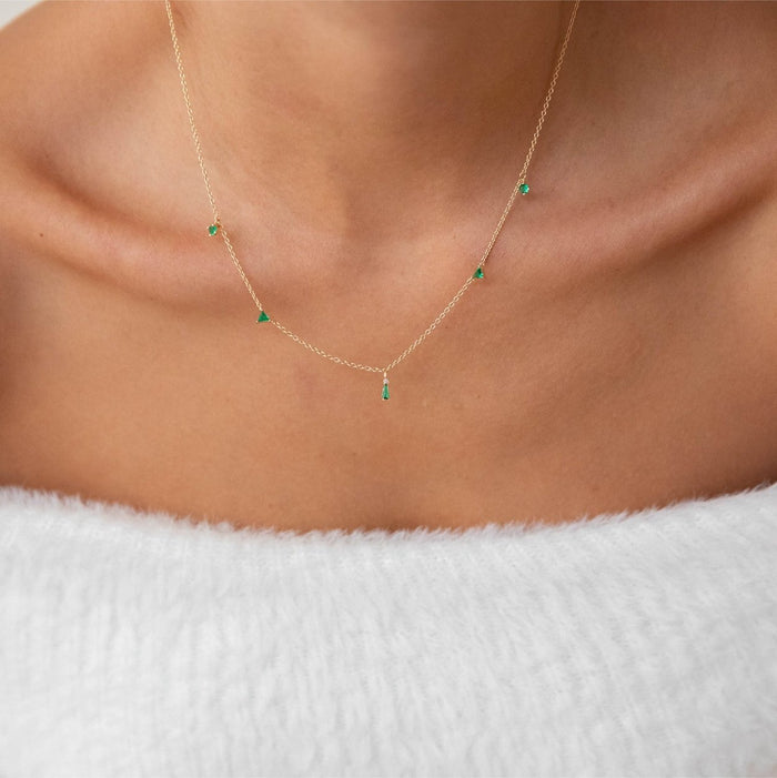 A close-up of a person’s neck wearing the Ivy Necklace by LLUME Jewelry—a gold plated piece with small emerald green gemstone charms, styled above a white textured off-the-shoulder top, perfect for dainty jewelry lovers.