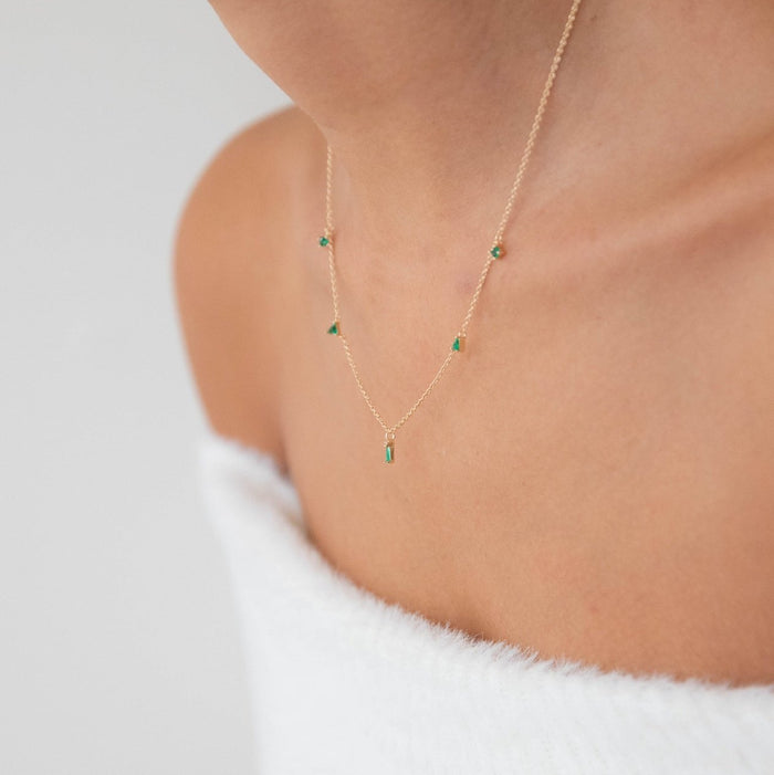 A person wearing an off-shoulder white fuzzy top and the Ivy Necklace by LLUME Jewelry, a dainty gold plated piece with small emerald green pendants, shown from the shoulders up against a plain background.