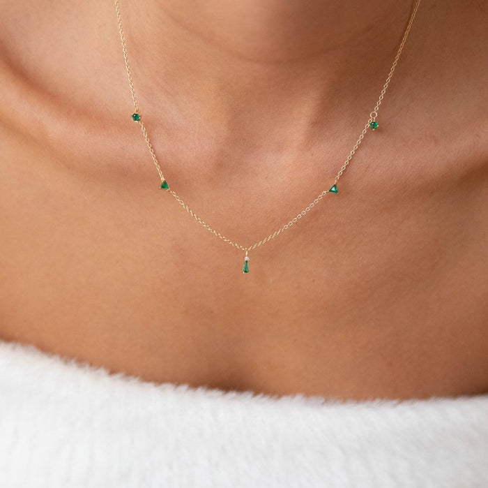 A close-up of a woman's neck wearing the Ivy Necklace by LLUME Jewelry, a dainty gold plated piece featuring five small emerald green pendants, styled over a white fuzzy off-shoulder garment.
