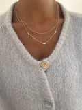 A person wears a soft, light gray knit cardigan with large buttons, styled with LLUME Jewelry's Juliet Daily Set—two delicate gold necklaces featuring small star-shaped pendants—highlighting the everyday jewelry and sweater texture.
