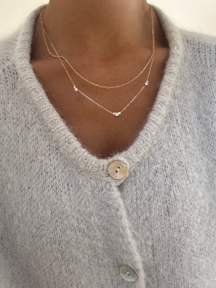 A person wears a soft, light gray knit cardigan with large buttons, styled with LLUME Jewelry's Juliet Daily Set—two delicate gold necklaces featuring small star-shaped pendants—highlighting the everyday jewelry and sweater texture.
