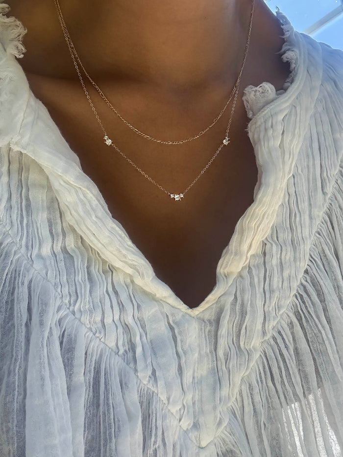 A person wears the Juliet Daily Set by LLUME Jewelry: a light, textured V-neck blouse pairs with two gold chain necklaces, each featuring flower pendants. Perfect everyday jewelry shown in soft, natural lighting.