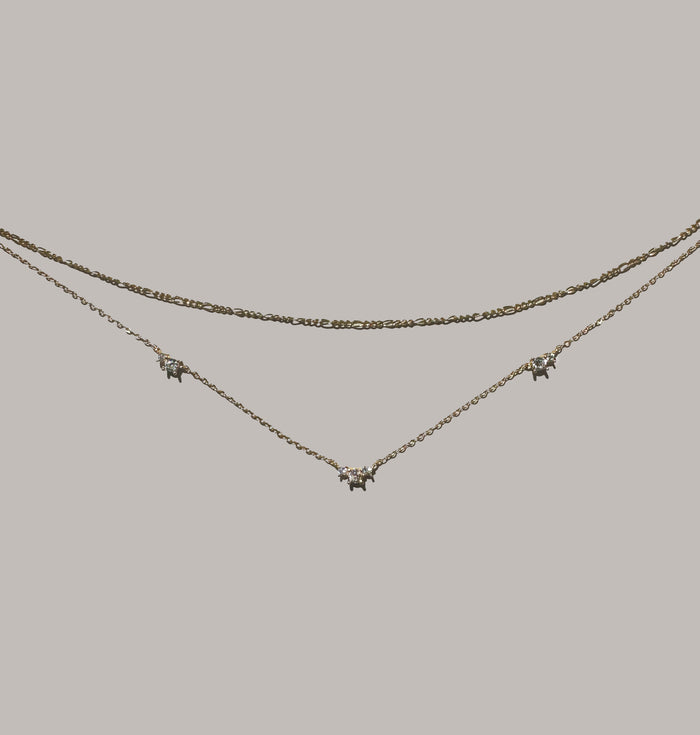 The Juliet Daily Set by LLUME Jewelry features two delicate gold necklaces—a slim-chain Juliet Necklace and the Colette Necklace with three star-shaped, gemstone-detailed pendants—perfect everyday pieces displayed on a plain light background.