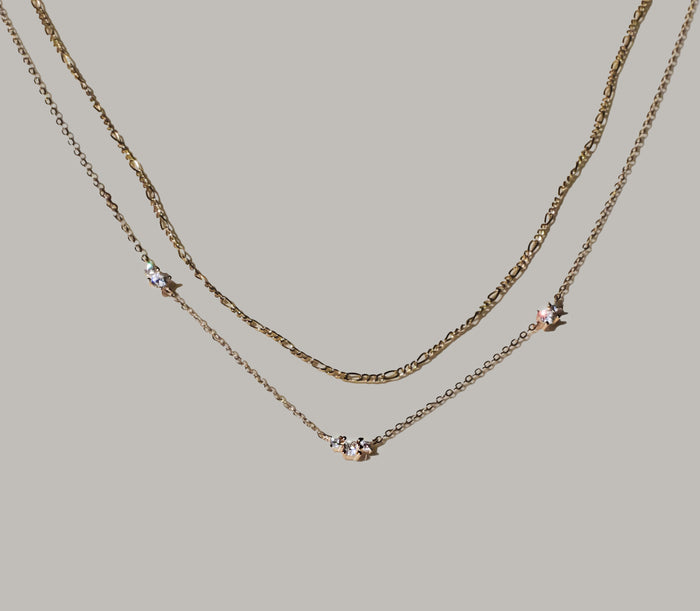 The Juliet Daily Set by LLUME Jewelry features two delicate gold necklaces—one simple and one with three sparkling gemstones—resting on a light gray background. This versatile set is perfect for stacking as your go-to everyday jewelry.