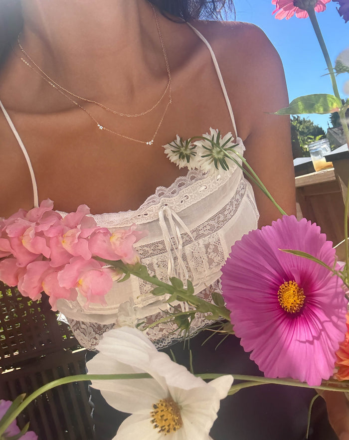 Wearing a white lace camisole and the Juliet Daily Set by LLUME Jewelry, featuring delicate gold pieces, she is surrounded by colorful flowers with sunlight streaming in the background.