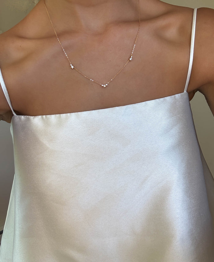 A person wearing a silky white spaghetti strap top and the LLUME Jewelry Juliet Necklace, an elegant gold-plated piece with small spaced beads, shown from the collarbone to mid-torso.