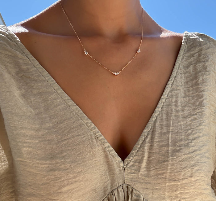 Outdoors beneath a clear blue sky, someone in a light beige v-neck blouse highlights the Juliet Necklace by LLUME Jewelry—an elegant gold-plated piece adorned with delicate star-shaped charms.