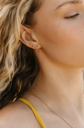 Close-up of a woman with wavy blonde hair wearing the LLUME Jewelry June-Prosecco Set—featuring a small heart and vertical bar earring—and a thin gold necklace. She wears a yellow top, her elegant earrings subtly highlighted.