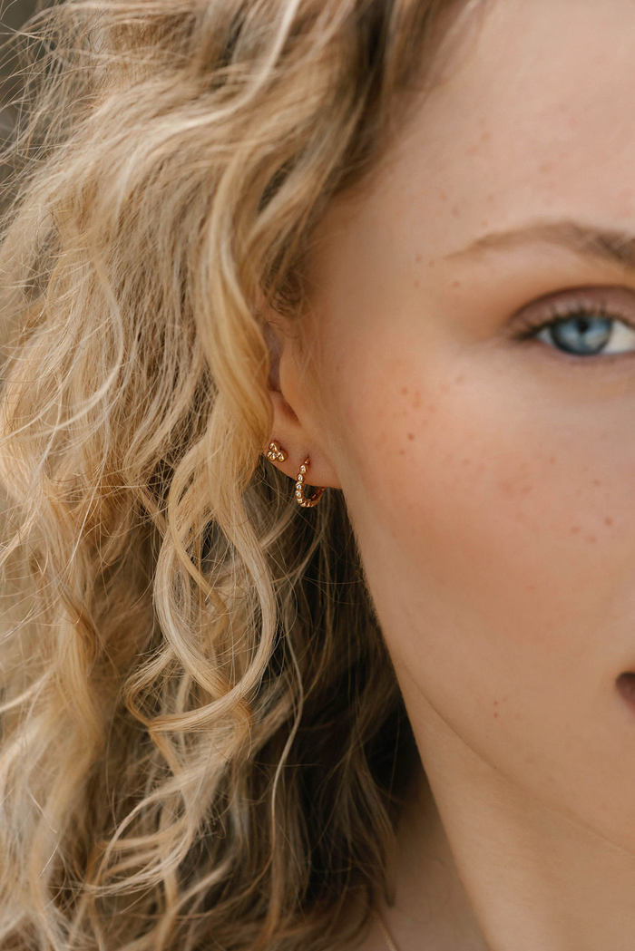 Close-up of a woman with blonde curls, blue eye, and light freckles, wearing the delicate June-Prosecco Set earring from LLUME Jewelry. The background is softly blurred.