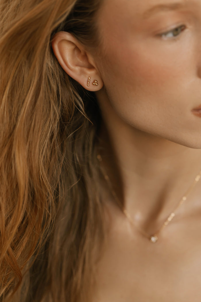 A close-up of a woman with wavy light brown hair wearing the LLUME Jewelry June Riley Stud Set—versatile gold stud earrings—and a delicate gold necklace with a small pendant, shown from the side in soft natural light.