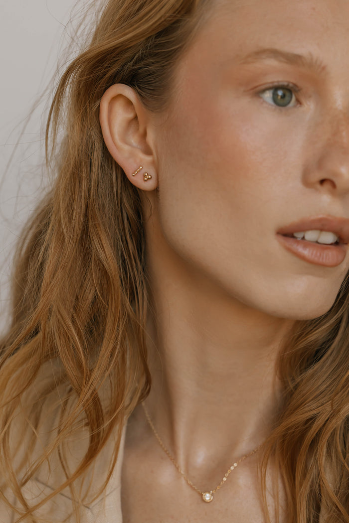 A close-up of a woman with wavy light brown hair, wearing gold stud earrings from the June Riley Stud Set by LLUME Jewelry and a delicate gold necklace. Her fair skin and neutral expression showcase her versatile jewelry style.