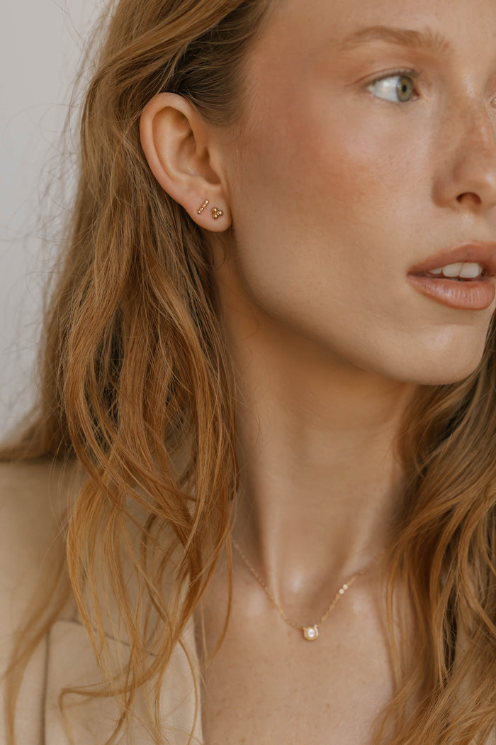 A woman with wavy light brown hair wears the June Riley Stud Set from LLUME Jewelry—versatile gold stud earrings and a delicate gold necklace. She looks right, her clear skin and natural makeup highlighted in soft lighting.