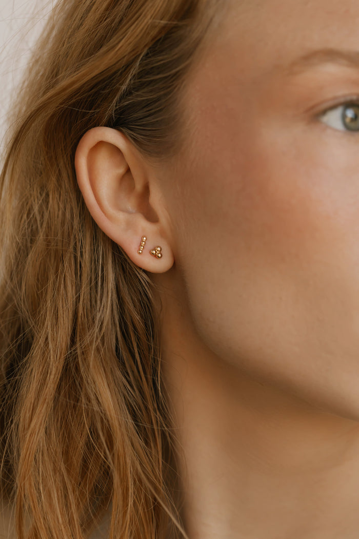 Close-up of a person's ear wearing the LLUME Jewelry June Riley Stud Set—gold earrings shaped like numbers 1 and 3 with small gemstones. This versatile set complements light brown hair and fair skin beautifully.
