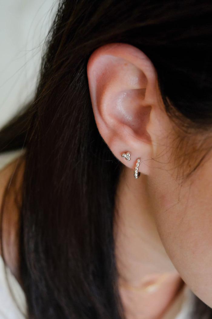 Close-up of a person's ear with minimalist gold-plated sterling silver earrings: LLUME Jewelry's small, heart-shaped June Studs and a small hoop with tiny stones. The person has straight, dark brown hair.