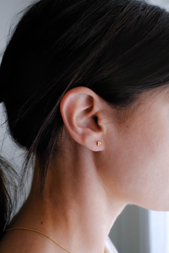 Close-up of a woman's ear wearing LLUME Jewelry's June Studs—minimalist gold plated sterling silver earrings. Her dark hair is pulled back, revealing her neck and jawline against a pale background.
