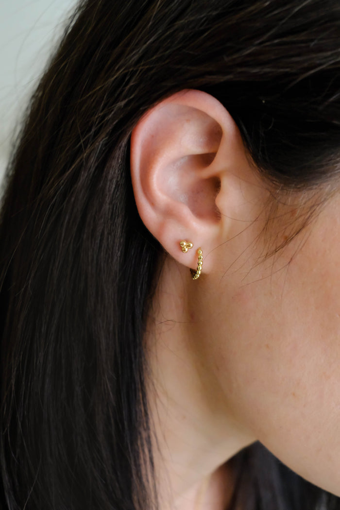 Close-up of a person with straight dark hair and light skin wearing minimalist earrings: LLUME Jewelry's June Studs and a small twisted hoop.