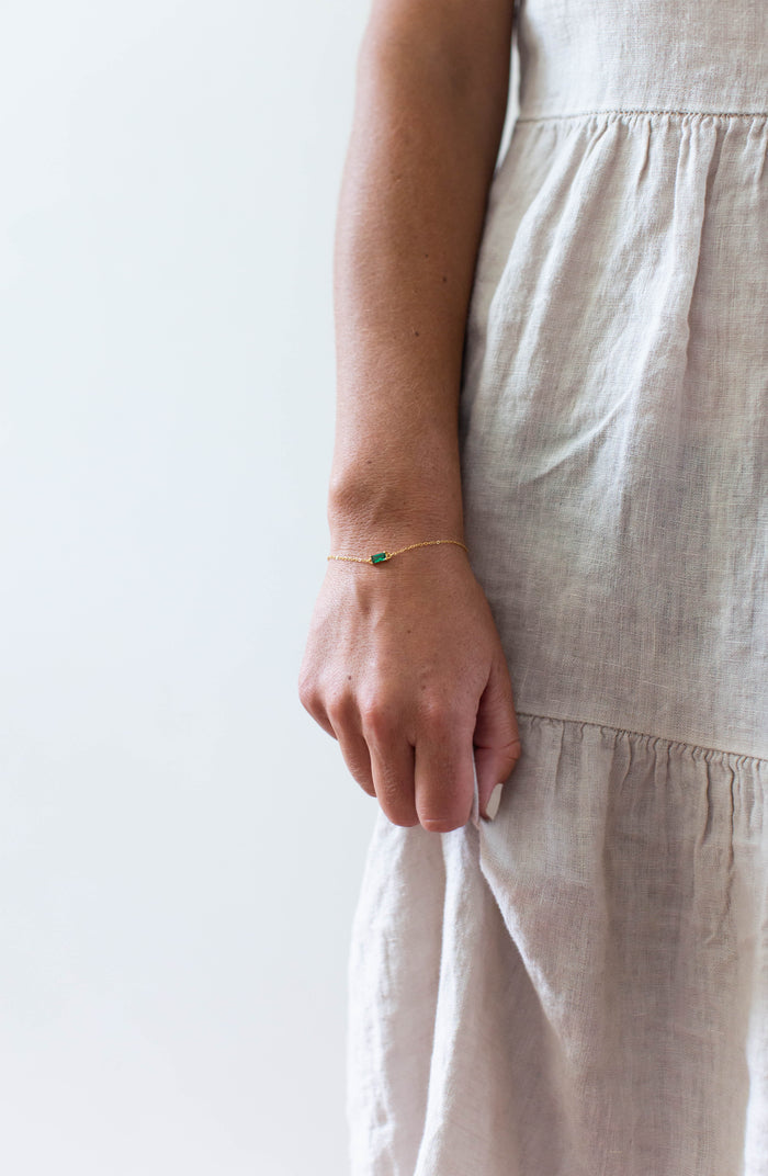 A person in a light beige dress stands with their arm relaxed, showcasing the LLUME Jewelry Kismet Bracelet—a dainty, adjustable gold-plated piece featuring a small emerald green stone—against a plain light background.