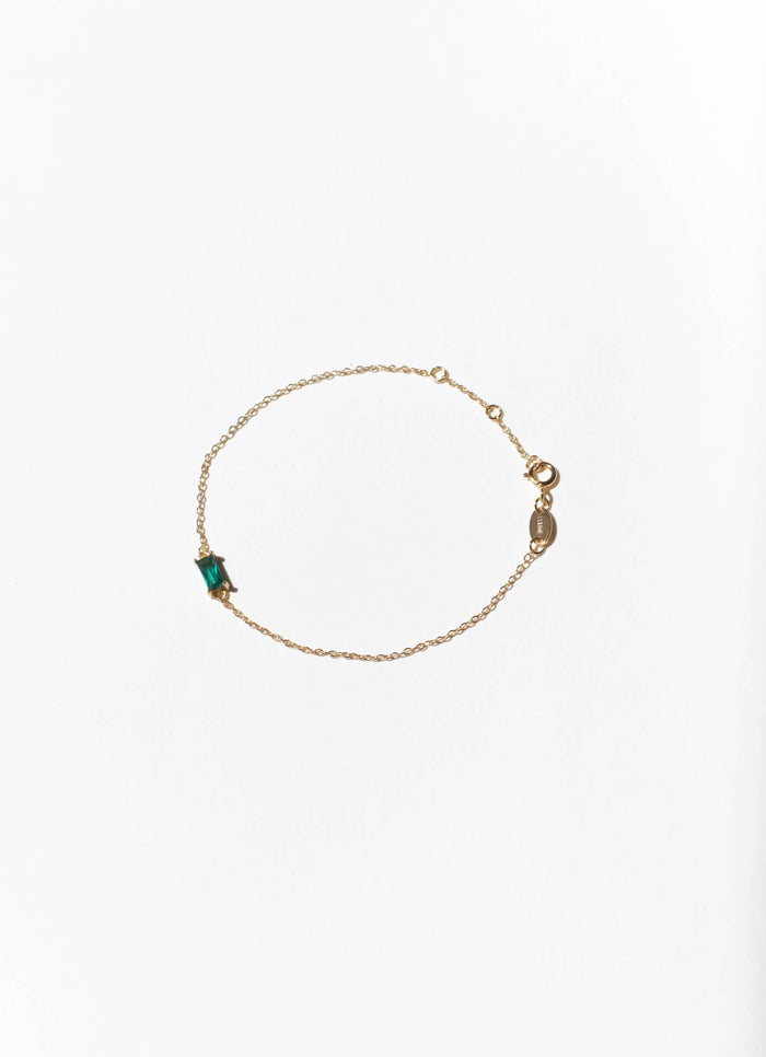 The Kismet Bracelet by LLUME Jewelry is a gold plated adjustable bracelet, featuring a small emerald green rectangular gemstone on one side and a lobster clasp with a round tag, shown on a plain white background.