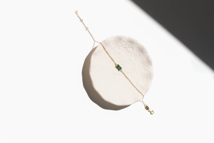 The Kismet Bracelet by LLUME Jewelry features a gold-plated chain and an emerald green rectangular gem centerpiece, displayed on a round white textured surface with dramatic lighting for a minimalistic look.