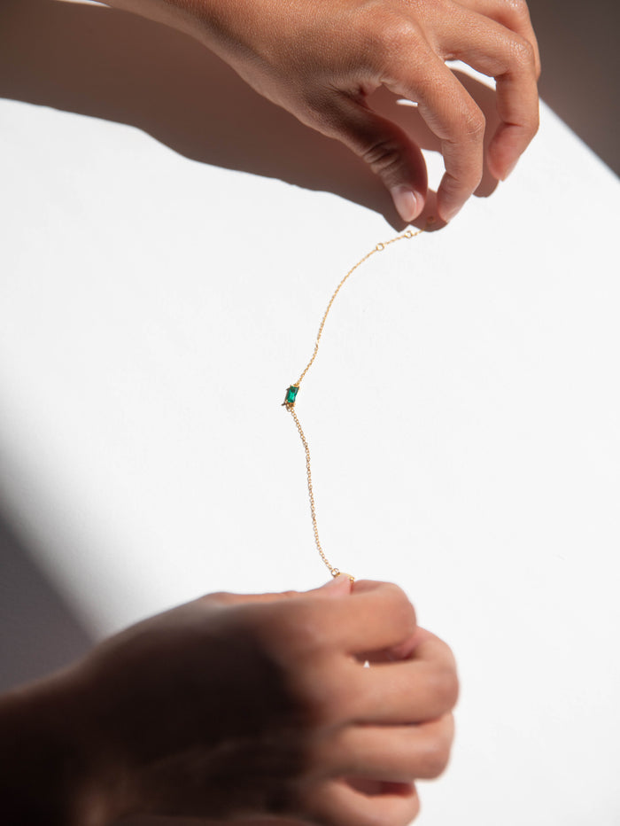 Two hands hold the Kismet Bracelet by LLUME Jewelry—an adjustable gold plated chain with an emerald green gemstone—casting soft shadows on a white surface under bright light.