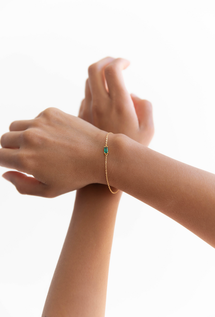 The Kismet Bracelet by LLUME Jewelry features a delicate gold-plated design with a small emerald green gemstone, shown worn on the wrist with both hands gently posed against a plain white background.