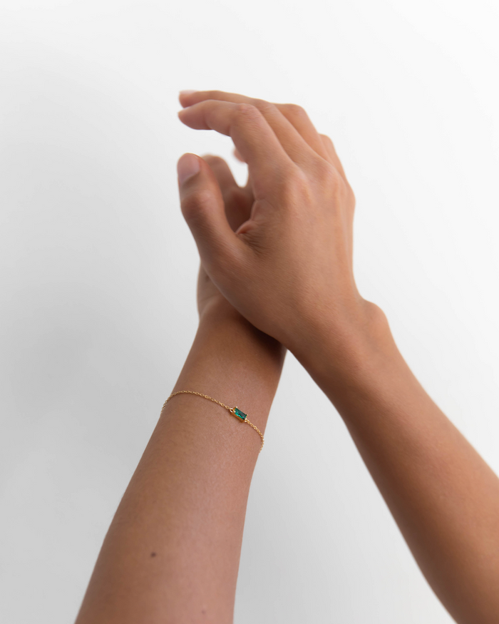 A close-up of two hands, one gently touching the other’s wrist, which is adorned with LLUME Jewelry’s Kismet Bracelet—an adjustable, dainty gold-plated piece featuring a small emerald green gemstone—set against a plain white background.