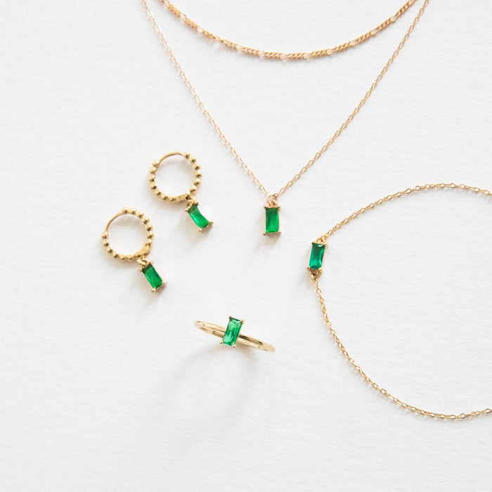 The Kismet Capsule by LLUME Jewelry features delicate gold jewelry with rectangular green gemstones—ideal for effortless layering. Set includes two necklaces, a bracelet, earrings, and a ring, elegantly arranged on a white surface.