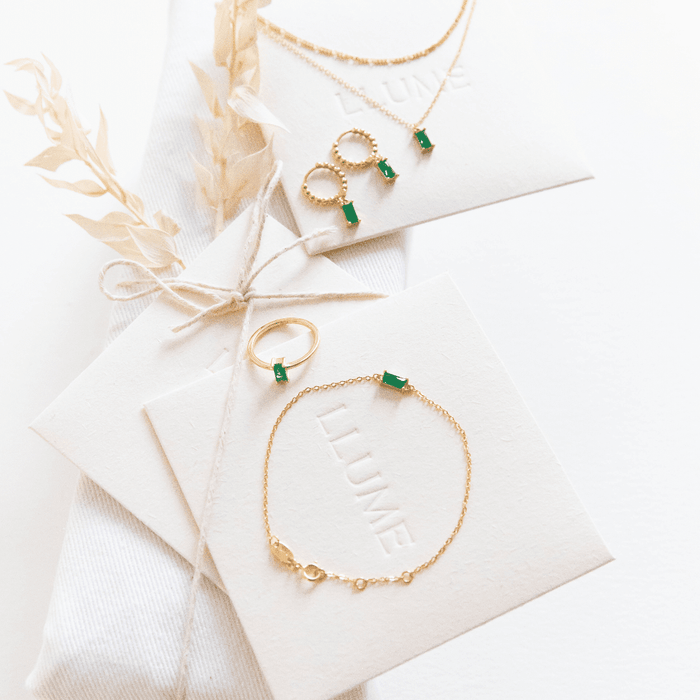 The Kismet Capsule jewelry set by LLUME Jewelry features gold pieces with green gemstones—necklace, earrings, ring, and bracelet—displayed on branded cards and perfect for effortless layering on a neutral background with beige dried flowers.