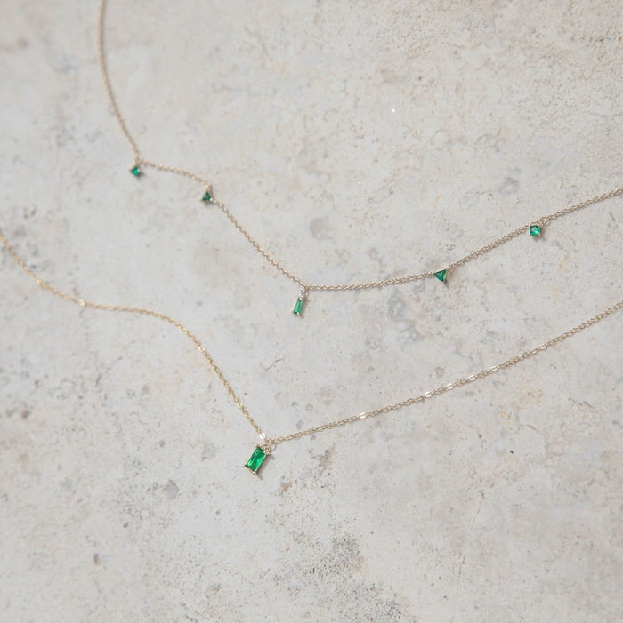 The Kismet Cluster necklaces by LLUME Jewelry feature gold chains with rectangular and triangular emerald green gemstones, evenly spaced and displayed on a light, textured stone surface.
