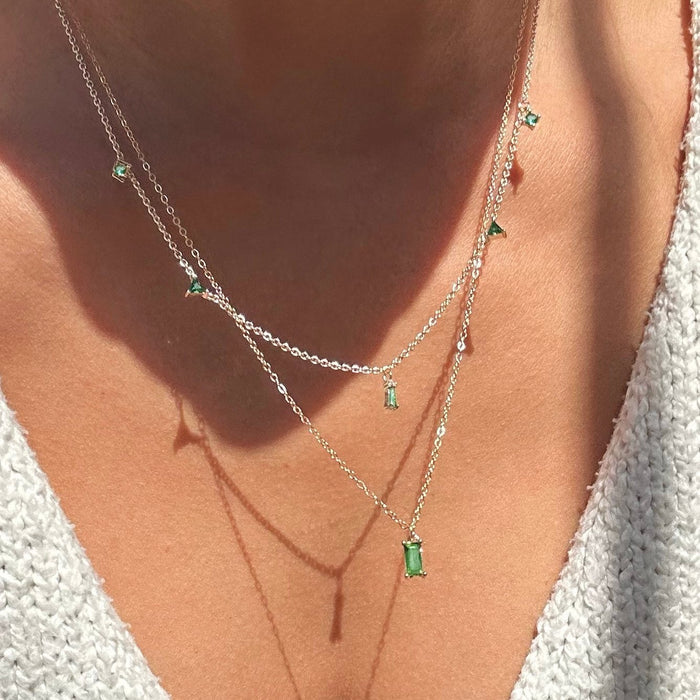Close-up of a person's neck wearing LLUME Jewelry's Kismet Cluster layered necklace—two delicate gold chains with small emerald green pendants. They wear a soft light-colored top, as sunlight casts gentle shadows on their skin.