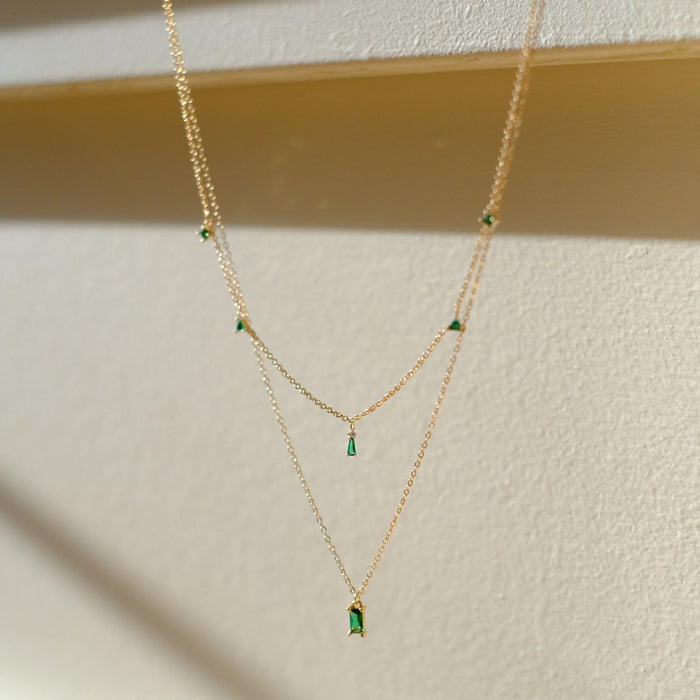 The Kismet Cluster layered necklace by LLUME Jewelry features small emerald green gemstone pendants spaced along a delicate chain, beautifully displayed against a light wall with soft sunlight and gentle shadows.