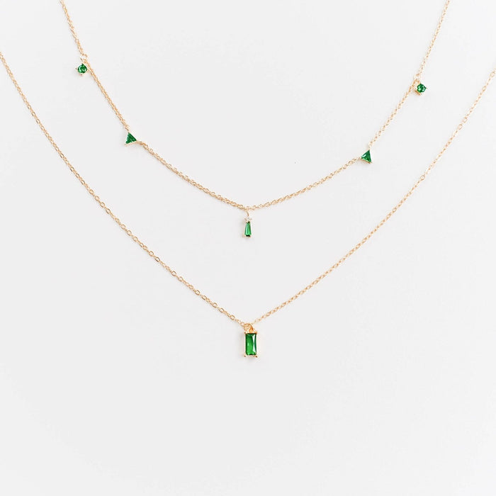 The Kismet Cluster necklace by LLUME Jewelry features delicate gold layers, each adorned with small emerald green gemstone pendants in unique shapes, creating a charming look against a white background.