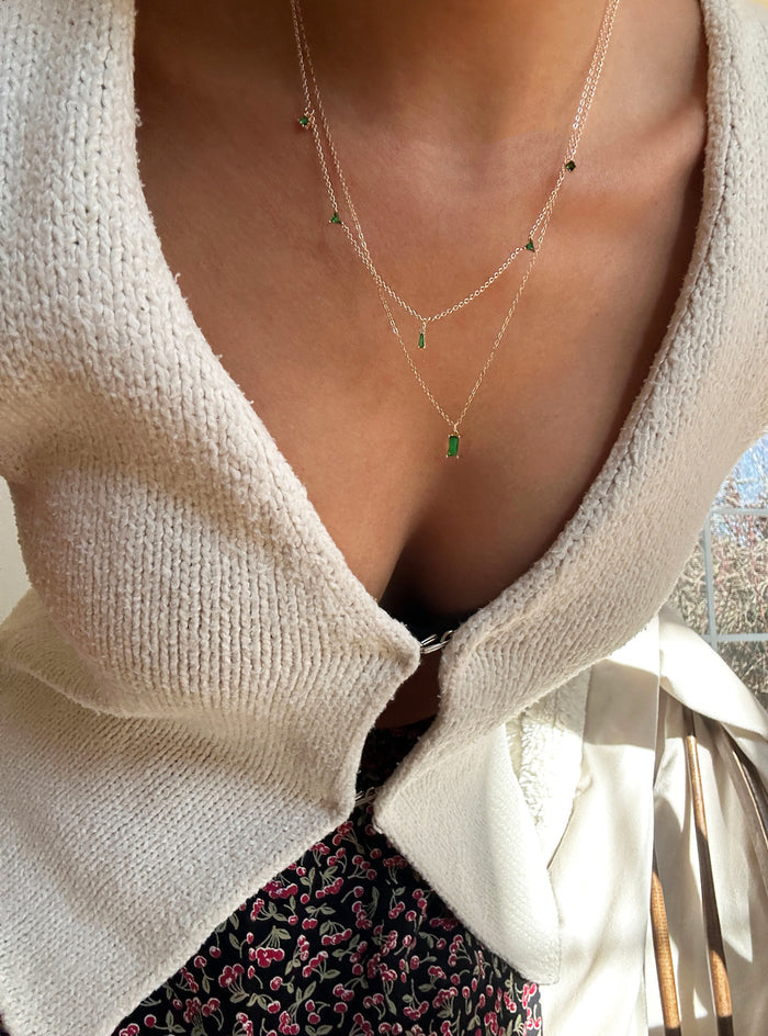 A person wears a cream-colored textured cardigan over a pink and green floral skirt, accessorized with the LLUME Jewelry Kismet Cluster necklace featuring small emerald green stones. The cardigan is slightly open in front.