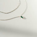 Two delicate silver necklaces from LLUME Jewelry’s Kismet Daily Set Silver rest on a white surface; one features a small rectangular green gemstone pendant, while the other is plain. Soft shadows are cast by the chains.