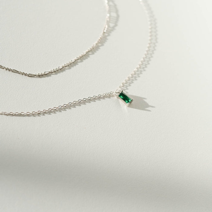 Two delicate silver necklaces from LLUME Jewelry’s Kismet Daily Set Silver rest on a white surface; one features a small rectangular green gemstone pendant, while the other is plain. Soft shadows are cast by the chains.