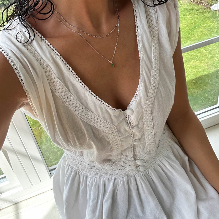 A woman in a white, textured, sleeveless dress with a deep V-neckline and buttons sits by a sunlit window. She layers two delicate necklaces—LLUME Jewelry's Kismet Daily Set Silver and another silver necklace. Her face is not visible.