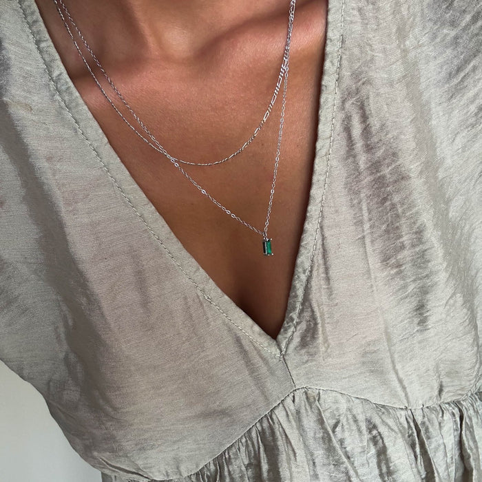 A close-up of a person with medium brown skin wearing a light, shimmery V-neck blouse and two delicate LLUME Jewelry silver necklaces, including the Kismet Daily Set Silver with a small green rectangular pendant.
