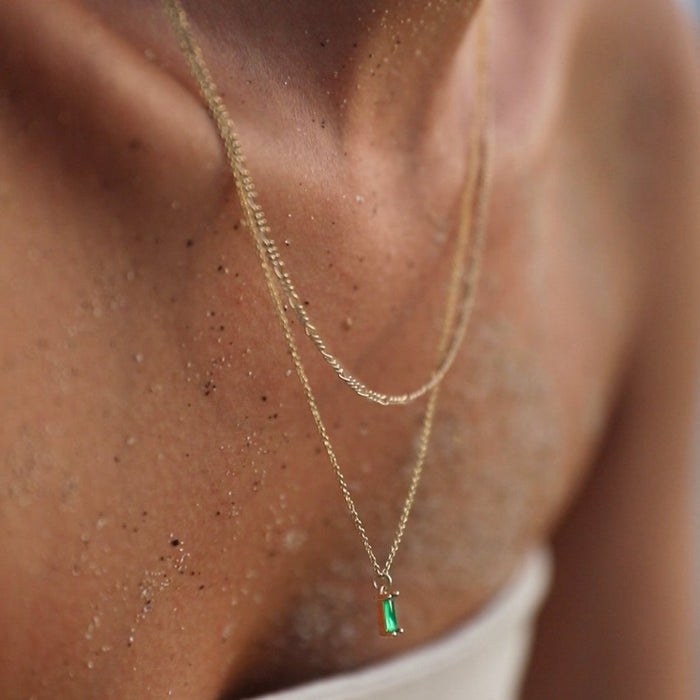 A close-up of a sandy-skinned person wearing the Kismet Daily Set by LLUME Jewelry, featuring layered necklaces with a small emerald green pendant, against a blurred neutral background.