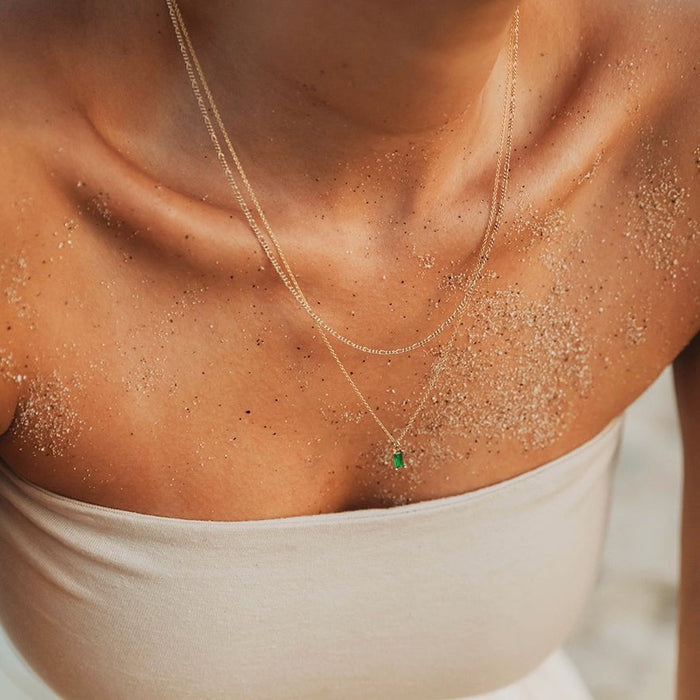 A close-up of a woman's sandy collarbone and shoulders, wearing the LLUME Jewelry Kismet Daily Set and another gold necklace with an emerald green pendant over a strapless beige top.