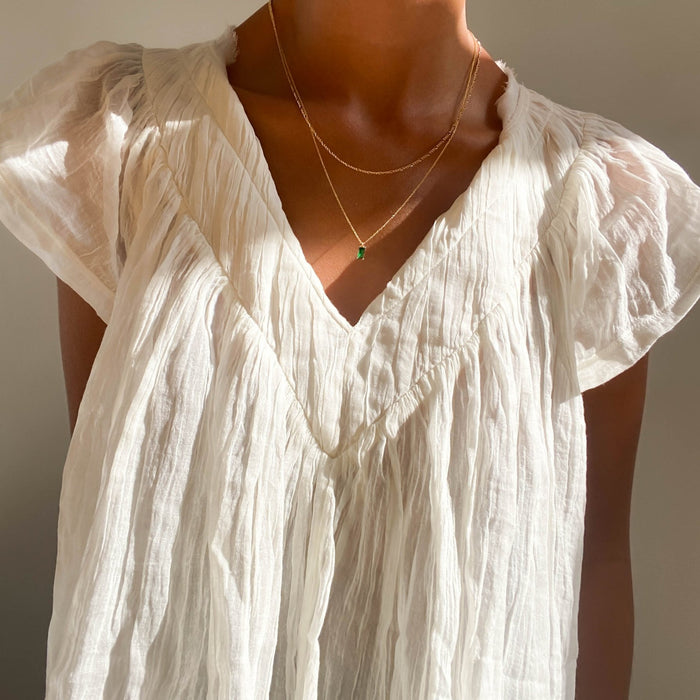 A person wearing a sheer, crinkled white blouse with short sleeves and a deep V-neck, accessorized with LLUME Jewelry’s Kismet Daily Set necklaces.