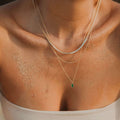 A close-up of a woman's sandy chest and collarbone, wearing the Kismet Favorites Set—layered chain necklaces with an emerald green pendant—paired with a strapless light-colored top.