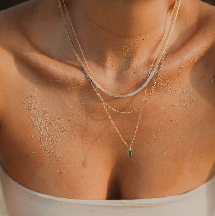 A close-up of a woman's sandy chest and collarbone, wearing the Kismet Favorites Set—layered chain necklaces with an emerald green pendant—paired with a strapless light-colored top.