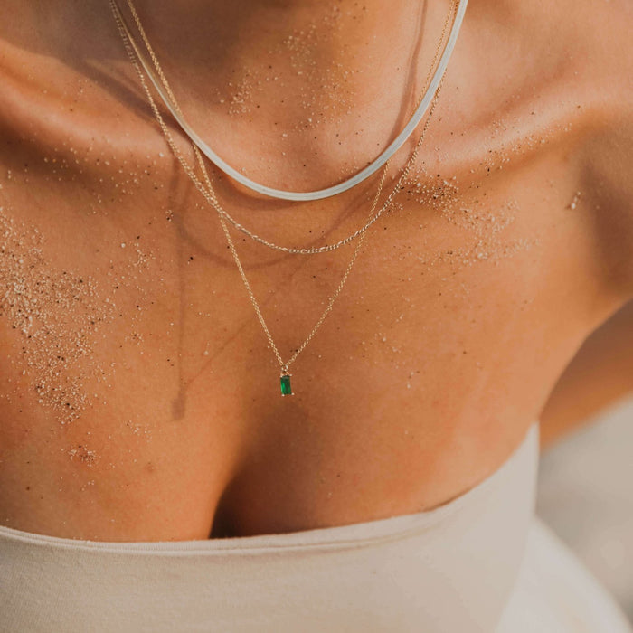 A close-up of a woman's sandy chest and collarbone, wearing a strapless top and the Kismet Favorites Set: a layered chain necklace set, with one necklace featuring an emerald green pendant.