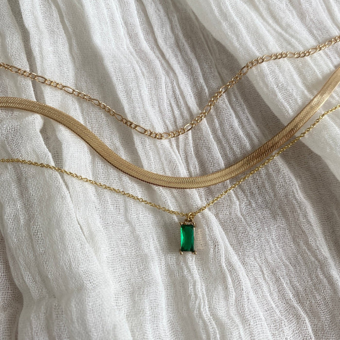 The Kismet Favorites Set features three gold layered necklaces on textured white fabric: one with an emerald green pendant and two plain gold chains of different styles and widths, creating a chic chain necklace set.