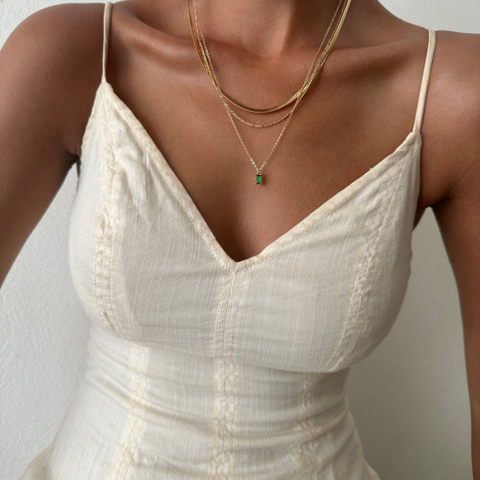 Wearing a cream sleeveless V-neck dress with thin straps, they accessorize with the Kismet Favorites Set: layered gold chain necklaces and a delicate emerald green pendant. The background is plain and light-colored.