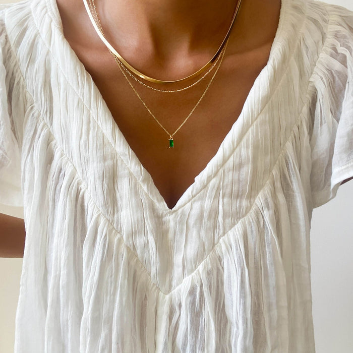 A person wearing a white, pleated V-neck blouse accessorized with the Kismet Favorites Set layered necklaces, featuring a chain necklace and one with an emerald green pendant.