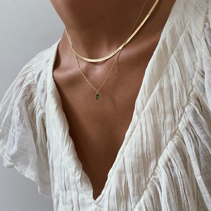 Wearing the Kismet Hailey Set—featuring a gold herringbone Skinny Hailey chain and emerald green Kismet Necklace—paired with a white, pleated v-neck blouse, delicate textures and jewelry shine under soft lighting.
