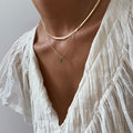Wearing the Kismet Hailey Set—featuring a gold herringbone Skinny Hailey chain and emerald green Kismet Necklace—paired with a white, pleated v-neck blouse, delicate textures and jewelry shine under soft lighting.